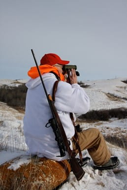 Man surveying with binoculars wearing a red ball cap, orange hood and white sweater with beige pants. The man has a rifle strapped to the back of his right shoulder. The sky is grey with rolling hills with patches of snow in the background.
