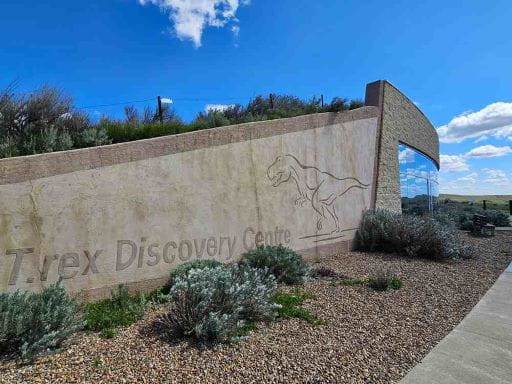 The T.rex Discovery Centre building located in Eastend SK. The image has Blue sky, sand, grey concrete wall with text engraved saying T-Rex discovery Center with a T-rex engraved in the wall.  